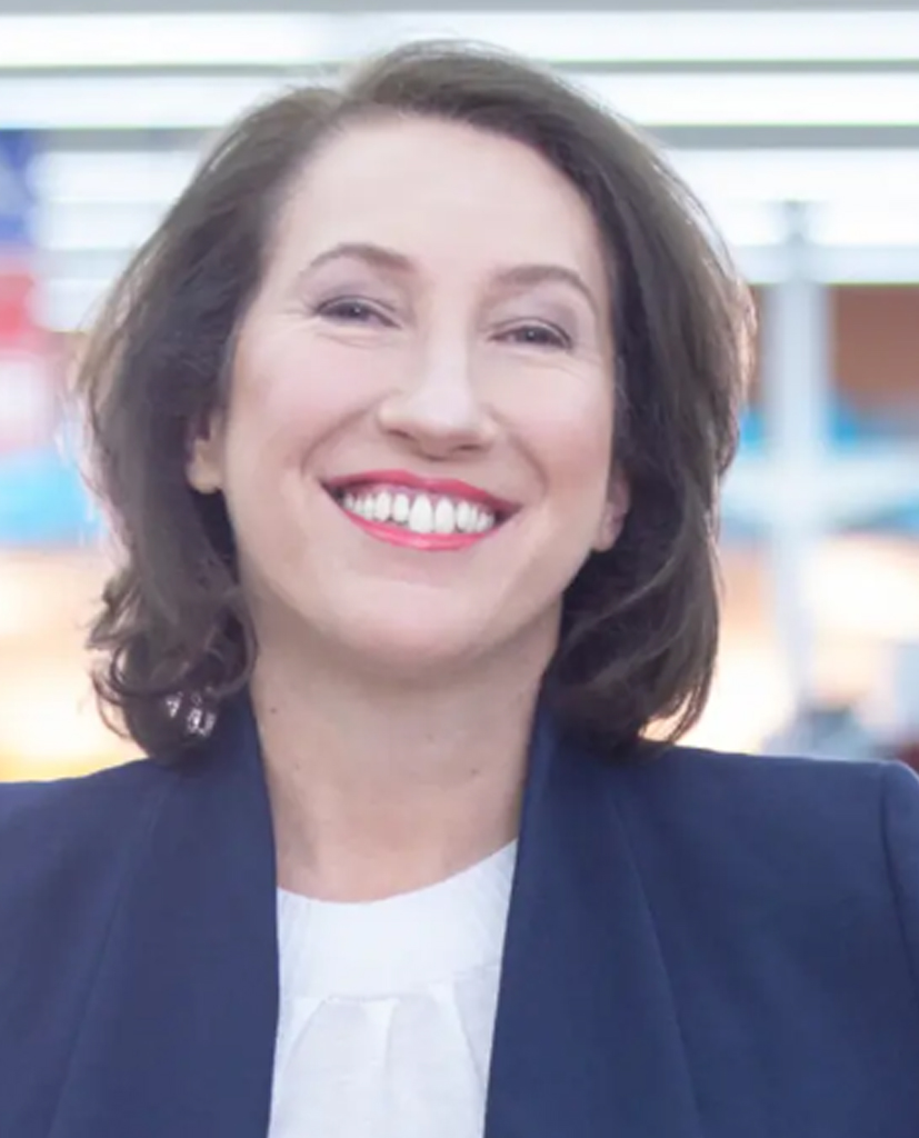 Porträt von Natalie Gerits in dunkelblauem Blazer und weißer Bluse, lächelnd in einem modern beleuchteten Marktumfeld.
